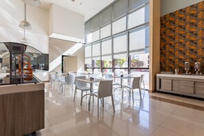 A modern cafe with a central counter, white chairs, and large windows offering views of the outside. - Apartamento no hotel Comfort (Taguatinga)