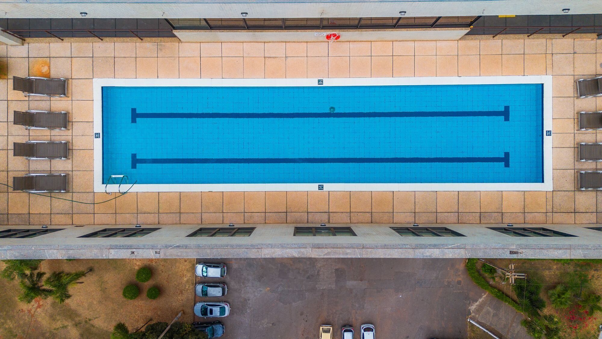 An aerial view of a swimming pool with a tiled deck, a covered area with seating, and a parking lot below.