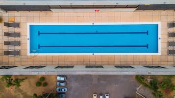 An aerial view of a swimming pool with a tiled deck, a covered area with seating, and a parking lot below.