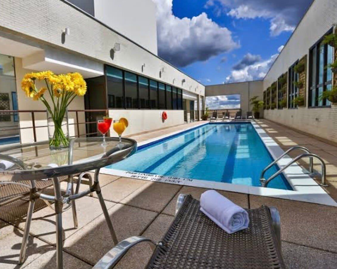 Una piscina con una mesa y sillas de vidrio, un jarrón con flores amarillas y un vaso de jugo de naranja.