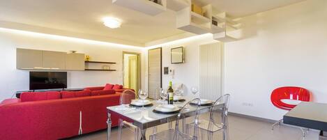 A modern living room with a red sofa, a dining table with clear chairs, and a kitchen area with white cabinets and a TV.