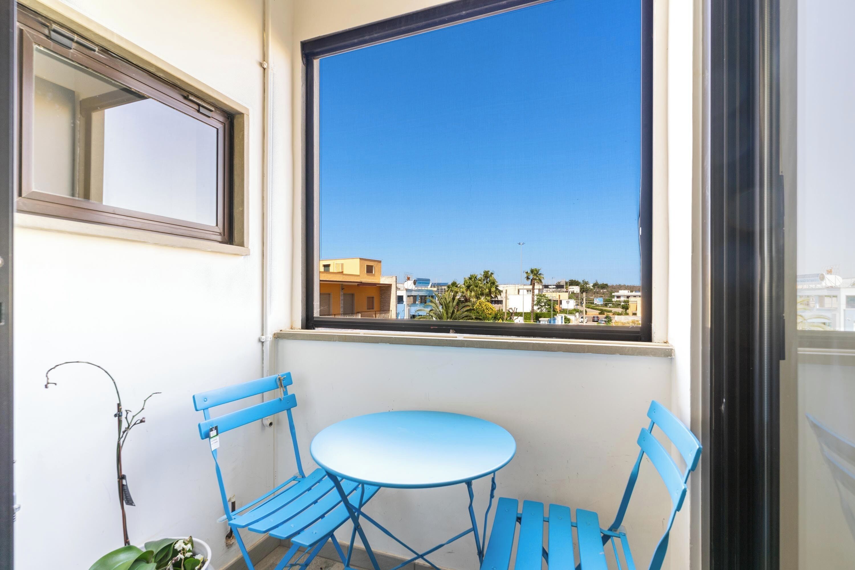 Ein Balkon mit blauem Tisch und Stühlen, einem Blumentopf und Blick auf Gebäude durch ein Fenster.