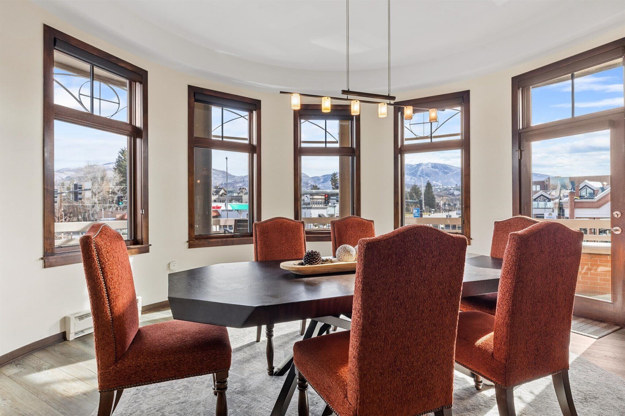 A dining area with a large table and six chairs, large windows with a view of mountains and a city, and a pendant light fixture.
