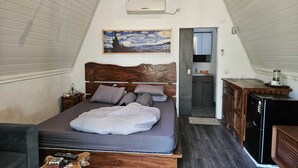 Una camera da letto con soffitto spiovente, un letto con testiera in legno, un quadro sopra il letto, un angolo cottura e una porta che conduce al bagno.