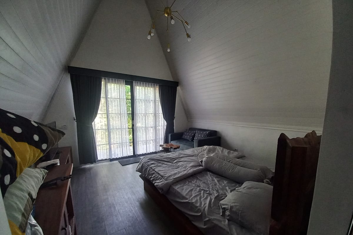 Une chambre avec un plafond en pente, un lit, une chaise, une petite table et une fenêtre avec des rideaux.