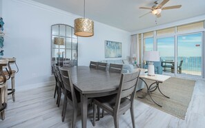 A dining area with a round table and chairs, a ceiling fan, and a view of the ocean through large windows. - Phoenix Orange Beach 804 3 Bedroom Condo by RedAwning (Orange Beach)