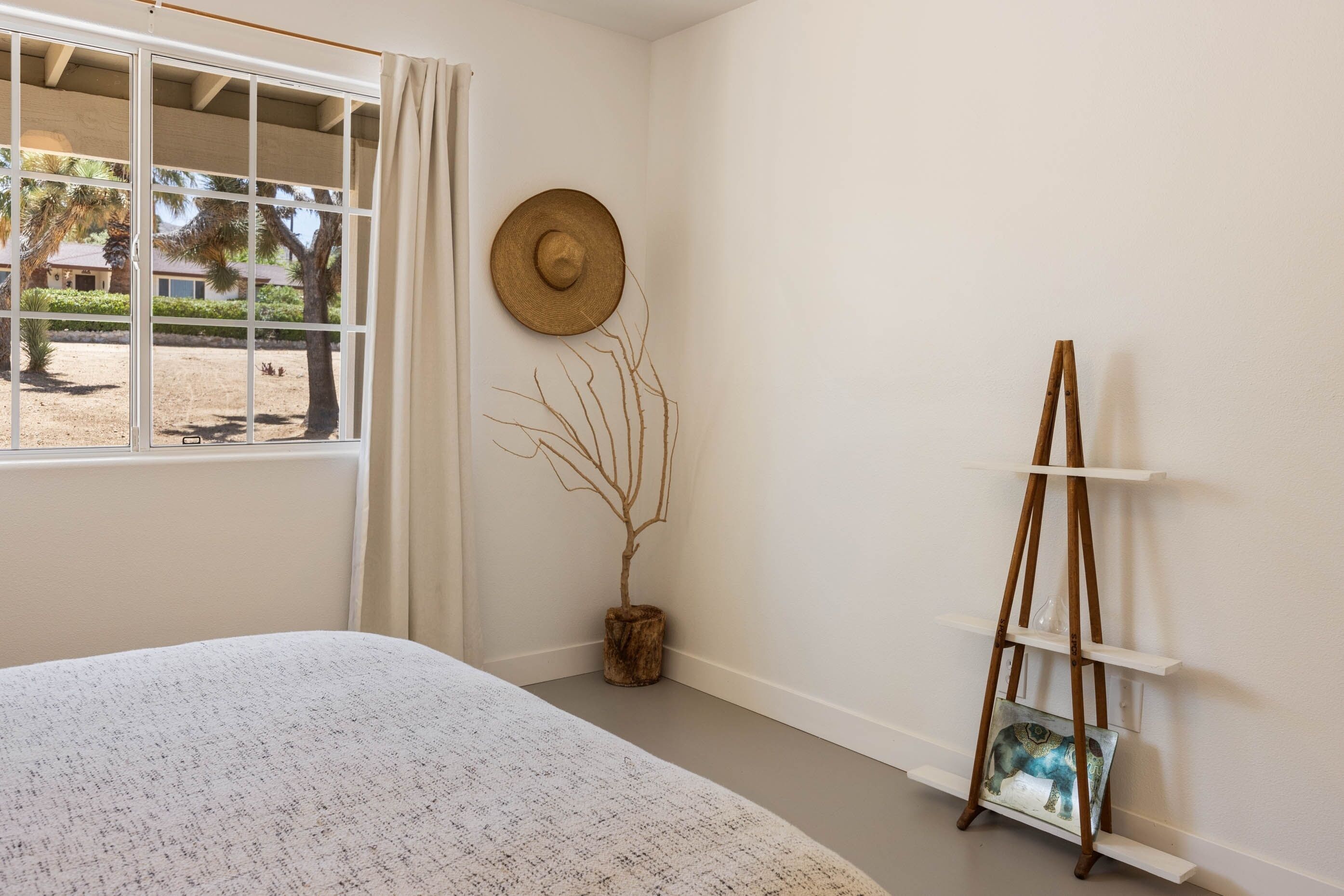 A bedroom with a large bed, a window with curtains, a wall-mounted hat, a small tree, and a wooden easel with a picture.