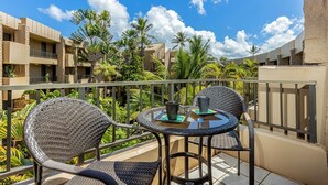 A balcony with wicker chairs and a small round table, surrounded by lush greenery and a clear blue sky. - Paki Maui 411 1 Bedroom Condo (Lahaina)