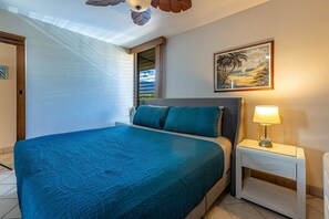 Ein Schlafzimmer mit einem Bett, einem Nachttisch, einer Lampe, einem Bild und einem Fenster mit Blick auf Palmen.
