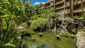 A lush garden with a pond, tropical plants, and a multi-story building in the background. - Paki Maui 411 1 Bedroom Condo (Lahaina)