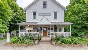 Un edificio blanco de dos pisos con un porche, señalado como "East Warren Schoolhouse". Un letrero indica "¡ABIERTO! ¡Todos son bienvenidos!"