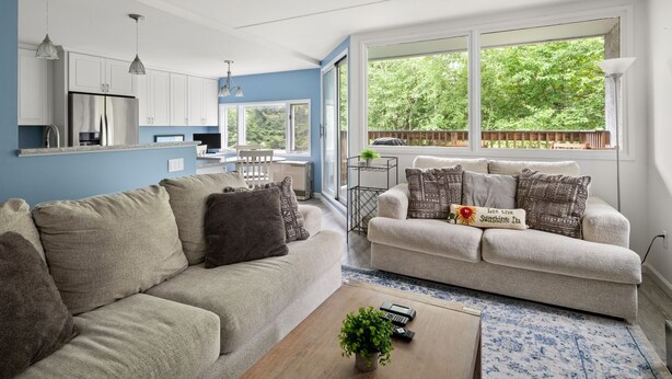 Una sala de estar moderna con un sofá beige, una mesa de centro con una planta, y vistas de la cocina y un espacio exterior a través de ventanas amplias.
