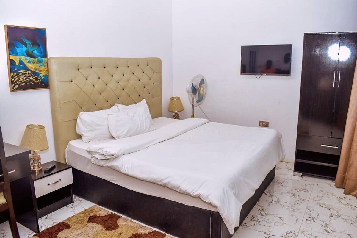 Un dormitorio ordenado con cabecera capitonada, cama con ropa blanca, mesita de noche con lámpara, televisor en la pared y armario.