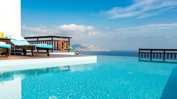 A clear blue infinity pool with lounge chairs and a wooden deck overlooking the sea.
