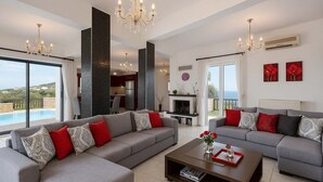 A spacious living room with two grey sofas, a coffee table, and a fireplace. There is a large window offering a view of the sea, and the room has a modern chandelier.