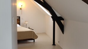 A narrow hallway with a sloped ceiling, a bed with a white headboard, a lamp, and a patterned rug on the floor. - Small house in the heart of a 15th century property. (Huillé-Lézigné)