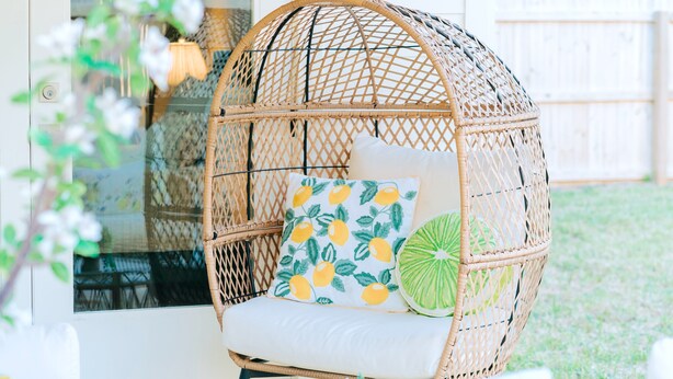 Un fauteuil suspendu en osier, doté d’un coussin et d’un oreiller à motifs d’agrumes, posé sur une surface blanche.