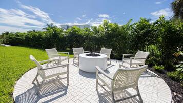A circular outdoor seating area with a fire pit, surrounded by white wicker chairs, on a paved patio with a view of a house and lush greenery.