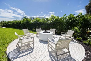 Un espace extérieur circulaire aménagé avec des sièges, doté d'un foyer, entouré de chaises en osier blanc, sur une terrasse pavée offrant une vue sur une maison et une végétation luxuriante.