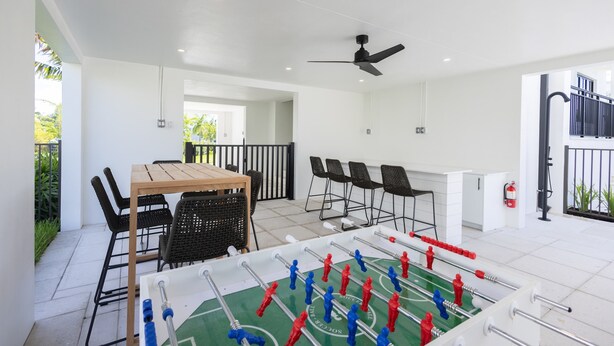 Una habitación con mesa de futbolín, una mesa de comedor con sillas, un ventilador de techo y vistas a un balcón con plantas.