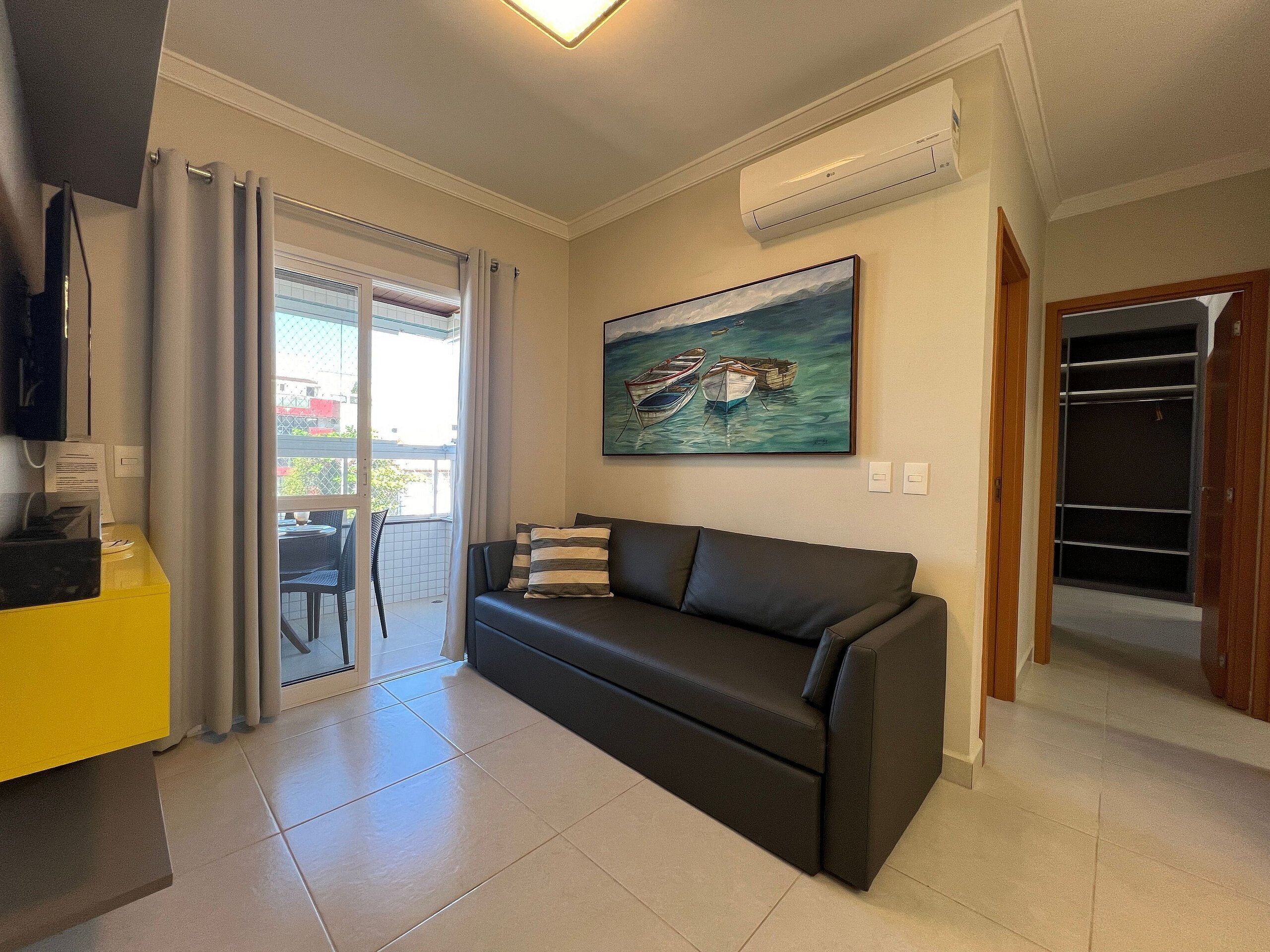 A modern living room with a black sofa, a painting of boats, a yellow cabinet, and a sliding glass door leading to a balcony.