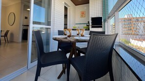 A dining area with a table set for two, surrounded by wicker chairs, a kitchen in the background, and a view through a window with a chain-link fence. - RESIDENCIAL VISTA VERDE (Ubatuba)