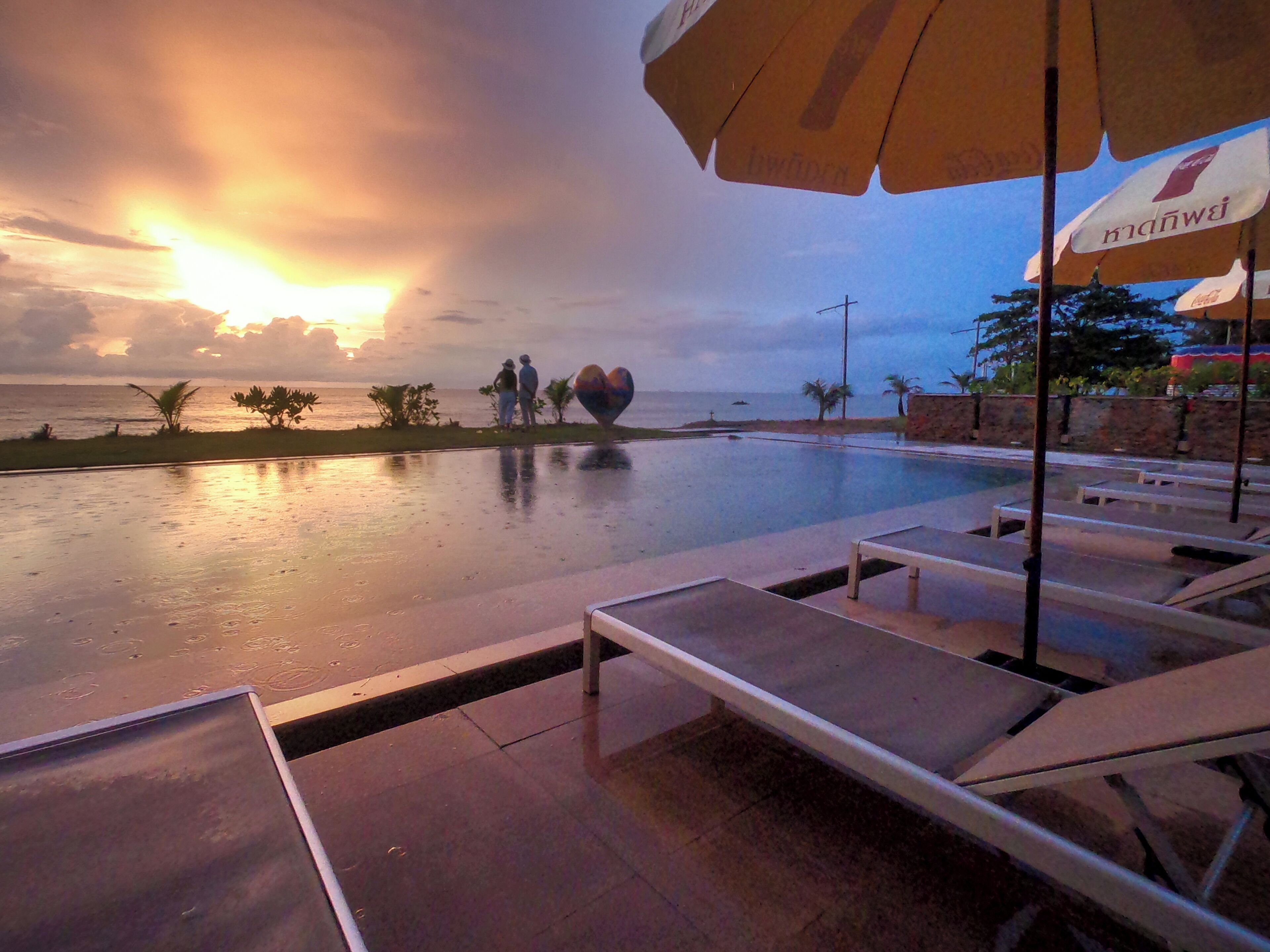 A poolside area with lounge chairs and umbrellas, a heart-shaped sculpture, and a view of the ocean at sunset.