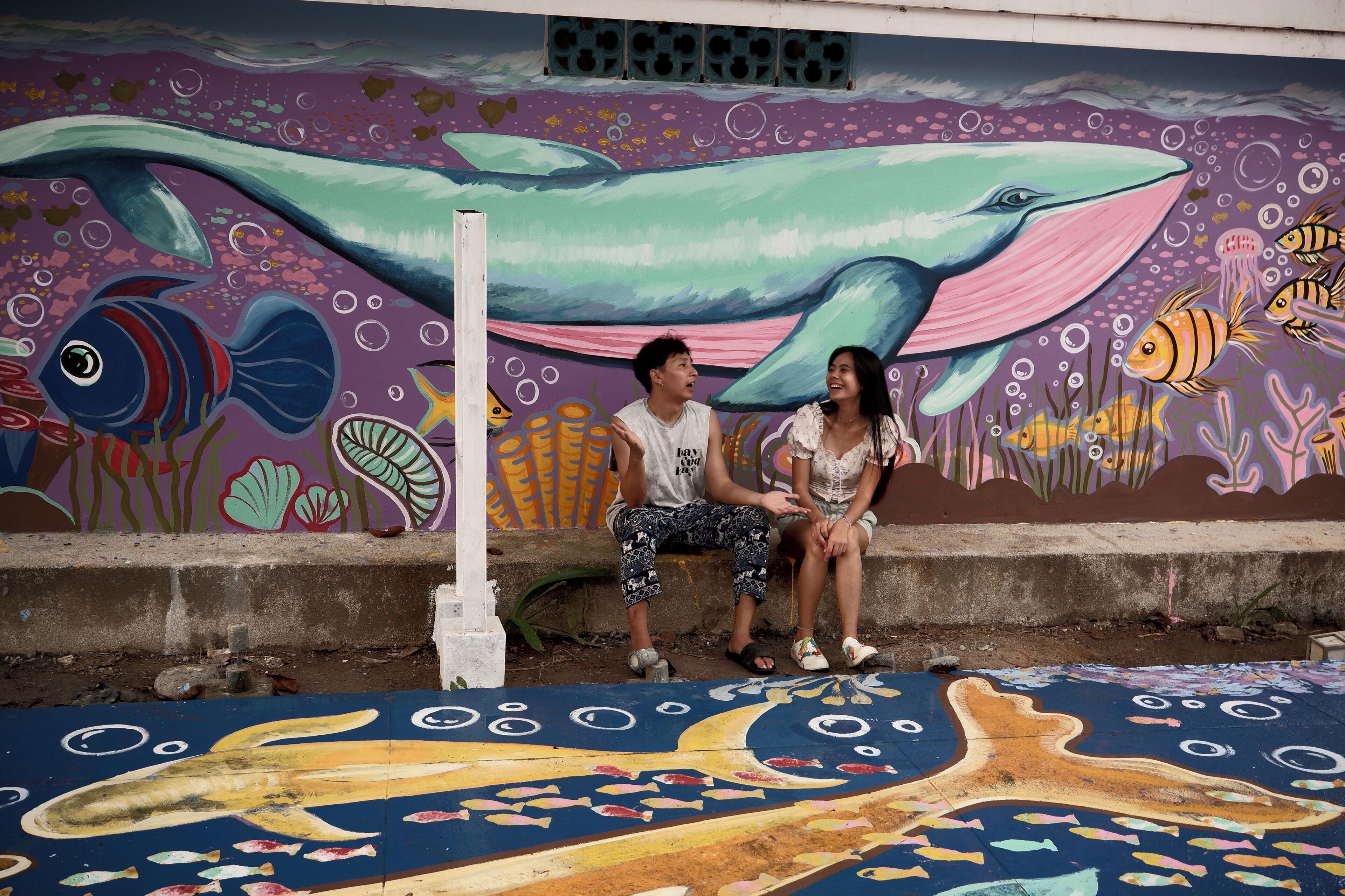 Deux personnes sont assises sur un banc devant une fresque colorée représentant une baleine et des poissons.
