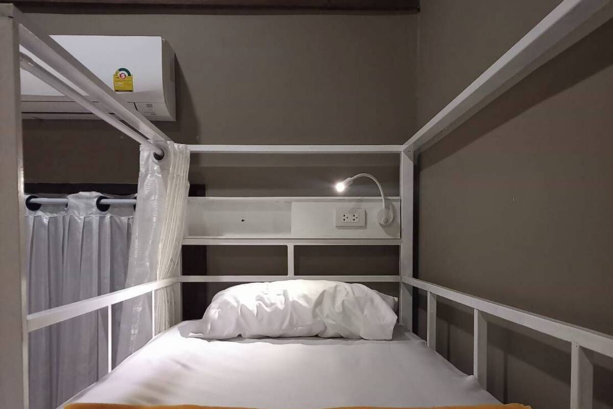 A bunk bed with a white mattress and a single pillow. There is a white lamp on the bed, and a power outlet is visible on the wall.