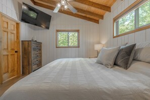 A bedroom with a large bed, wooden ceiling beams, a TV mounted on the wall, a window with a view of trees, and a dresser with drawers. - On The Rocks by Innsbrook Vacations! (Innsbrook)