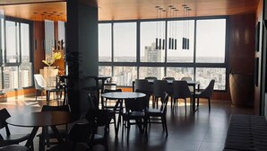 A spacious room with multiple tables and chairs, large windows offering a city view, and modern pendant lighting.