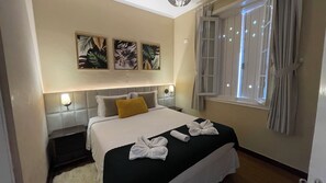 A hotel room with a bed, a nightstand, a wall-mounted lamp, and a window with curtains. - Vila Tio Lé (Campos do Jordão)