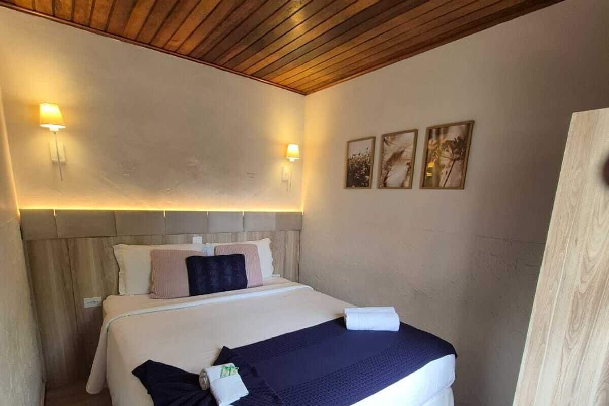 Un dormitorio con techo de madera, una cama con ropa de cama blanca y manta azul oscuro, dos lámparas de pared y tres cuadros en la pared.