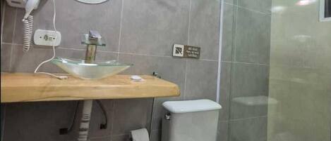 A bathroom with a glass shower enclosure, a white toilet, a glass sink with a mirror above it, and a hairdryer on the wall.