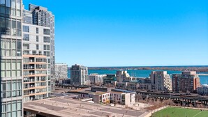 A high-rise building with a modern glass facade, a parking lot, and a view of a body of water and other buildings in the distance.