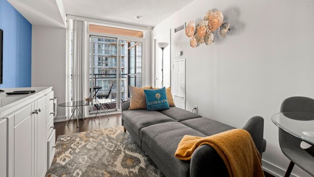 A modern apartment with a grey sofa, a small dining table, and a large window offering a view of buildings.