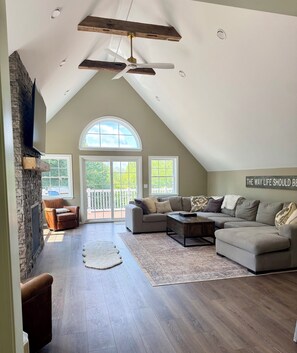A spacious living room with a high ceiling, a fireplace, a large sectional sofa, and a view through the windows. - Peaceful Maine Home with Mountain Views (Kingfield)