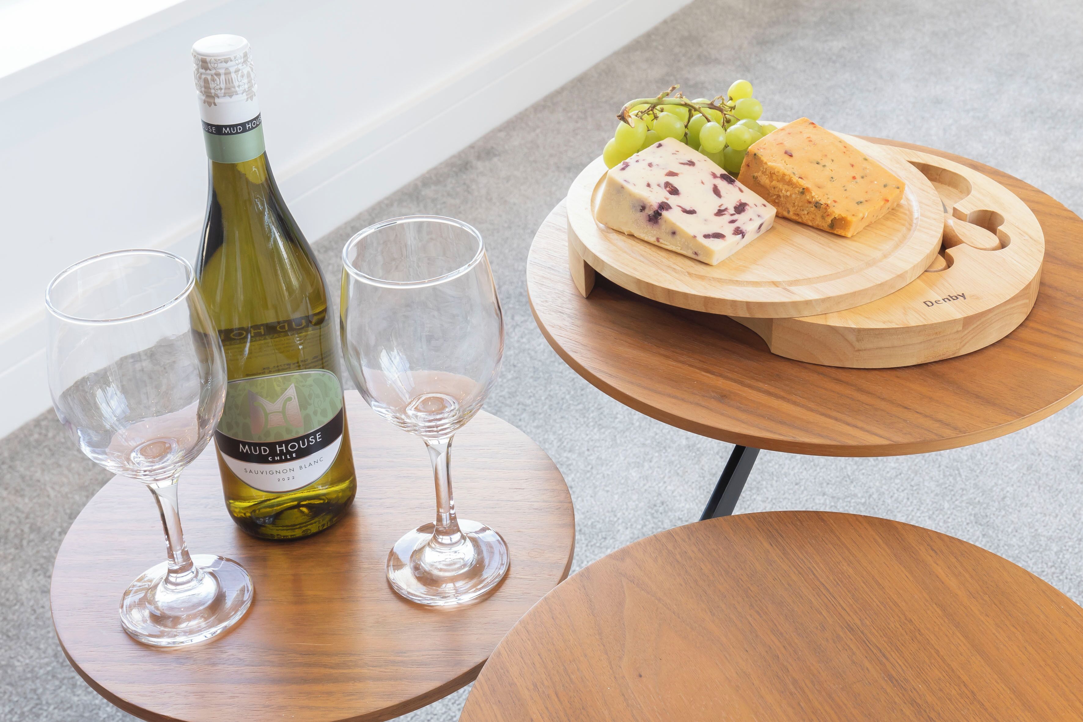 A bottle of wine, two glasses, and a wooden board with cheese and grapes on a round table.