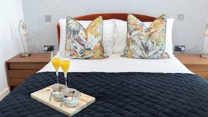 A neatly made bed with a black quilted bedspread, two colorful pillows, a wooden bedside table with a lamp, and a tray with jars and glasses of orange juice. - Elliot Oliver - The Penthouse 2 Bedroom Apartment With Parking (Cheltenham)