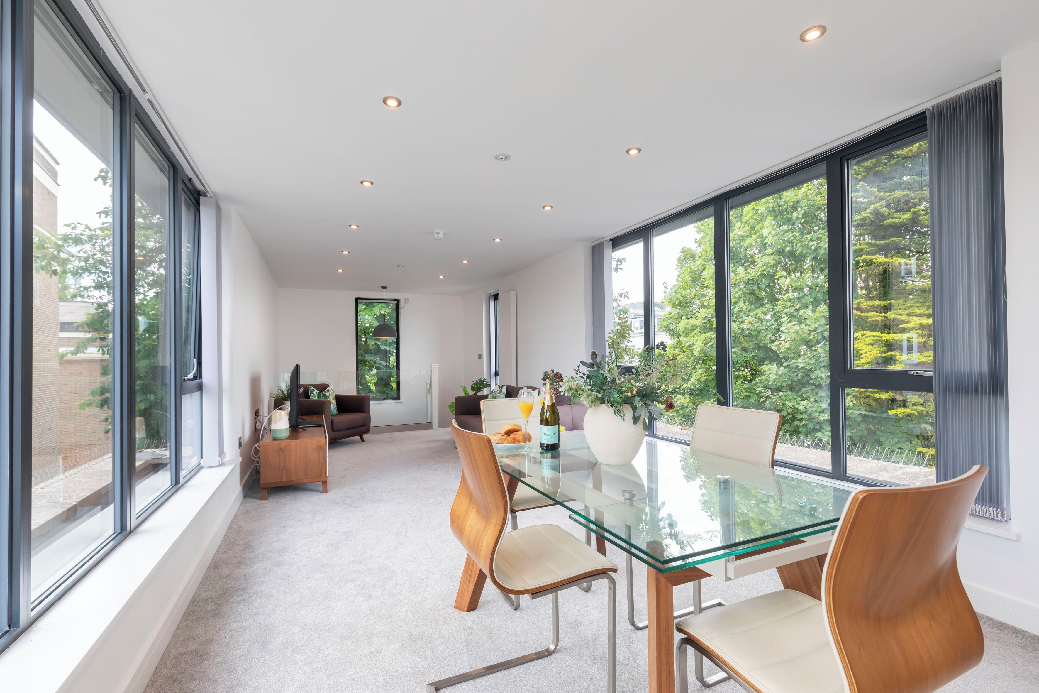 A modern dining area with a glass table, wooden chairs, and a view of greenery outside.