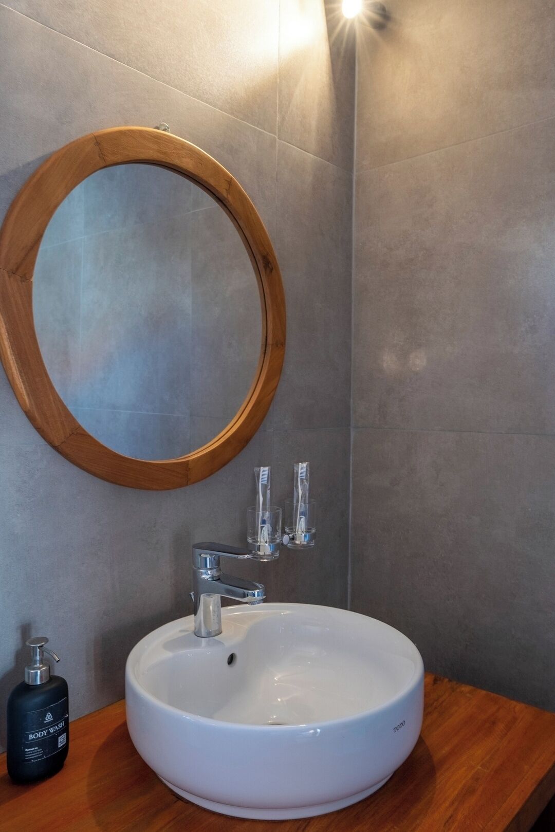 A bathroom with a round wooden-framed mirror, a white sink, and a bottle of body wash.