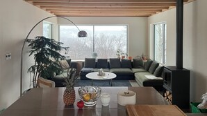 A modern living room with a wooden ceiling, a large window, a sofa set, a dining table with a pineapple and a bowl, and a fireplace. - Chalet des Sentiers - An architectural experience at Lac Fidèle, Entrelacs  (Entrelacs)