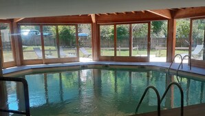 Una piscina interior con paredes de vidrio transparente, vigas de madera y suelo de baldosas.