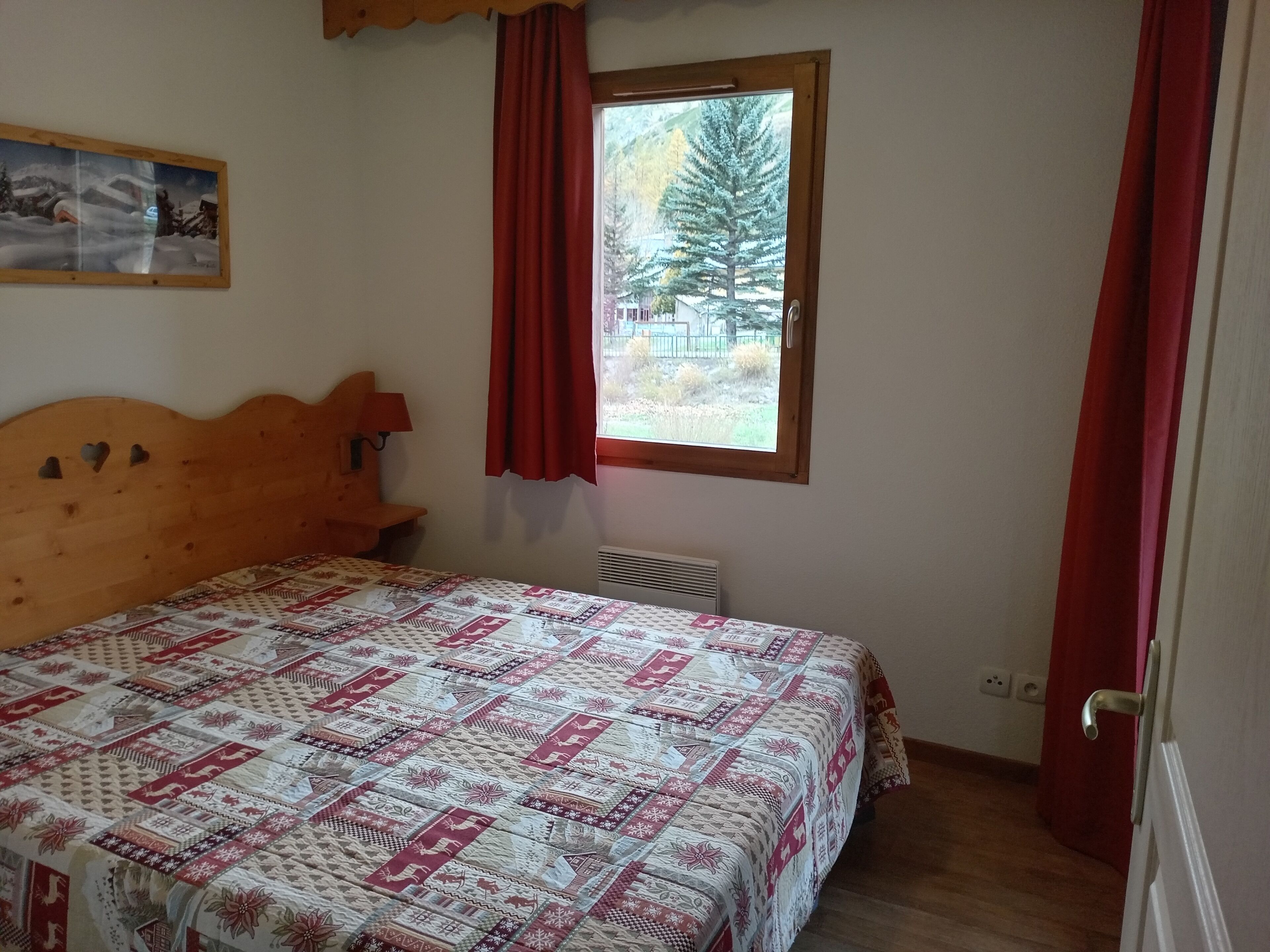 Ein Schlafzimmer mit einem Holz-Kopfteil, einer gemusterten Tagesdecke, einem Fenster mit roten Vorhängen und einem Bild an der Wand.