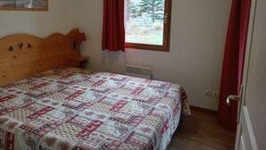 Un dormitorio con cabecera de madera, colcha estampada, ventana con cortinas rojas y un cuadro en la pared.