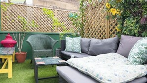 Eine Terrasse mit einem grünen Korbsessel, einem kleinen Tisch und einem roten Blumentopf.