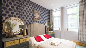 Ein Schlafzimmer mit Wand im geometrischen Muster, einem Bett mit roten Kissen, einem runden Spiegel und einer Kommode mit goldfarbenen Griffen.