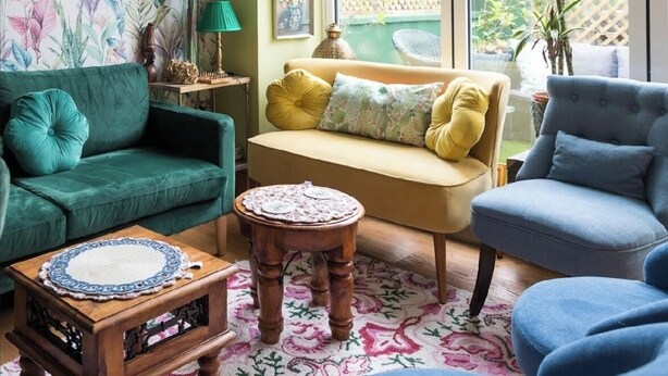 Ein gemütliches Wohnzimmer mit einem grünen Sofa, einem blauen Sessel und einem gelben Sofa. Ein Holztisch und ein kleiner Beistelltisch mit einer Lampe stehen im Raum. Das Zimmer hat ein großes Fenster mit Blick auf einen Garten.