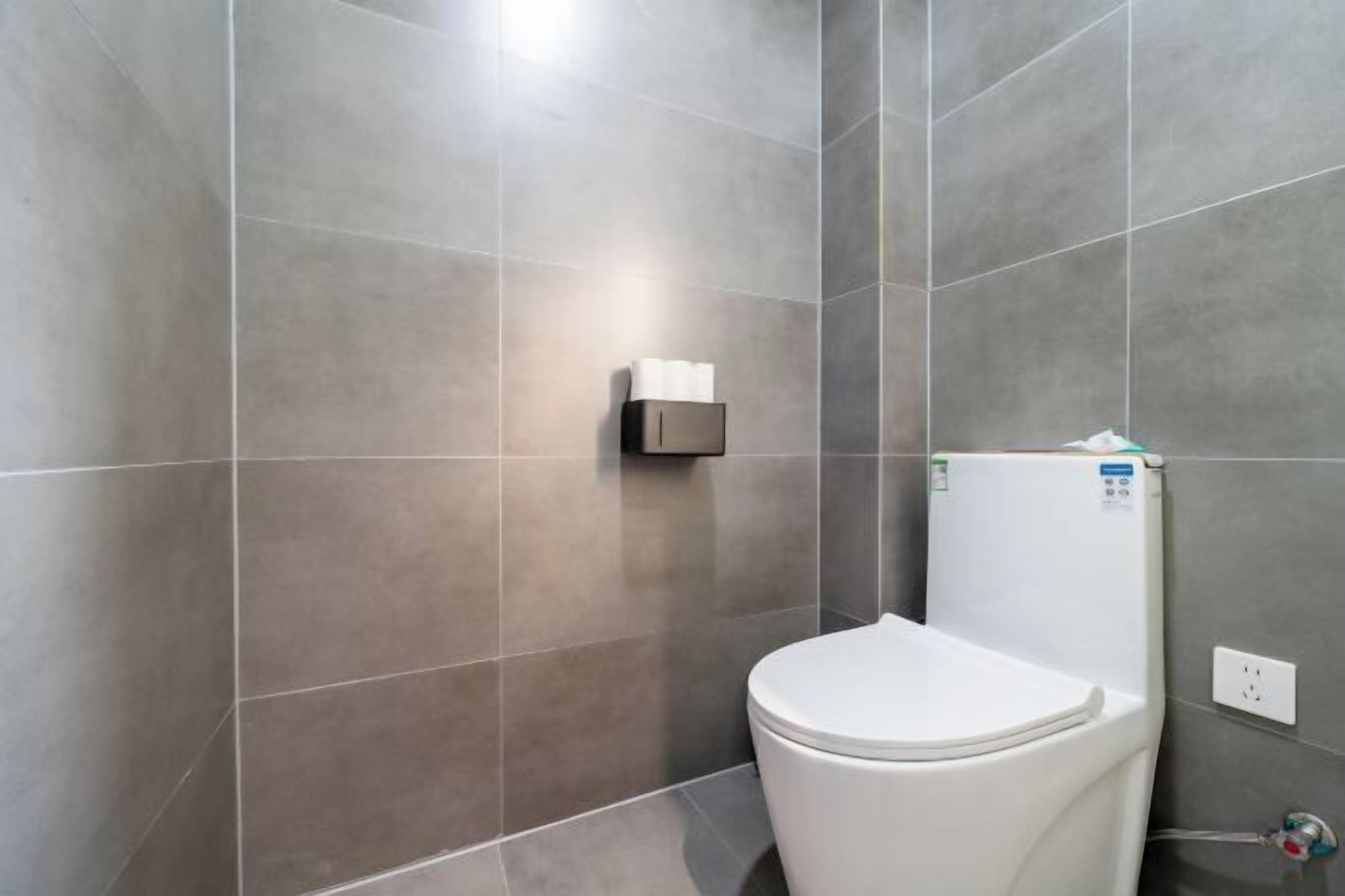 A modern bathroom with grey tiles, a white toilet, and a towel dispenser.