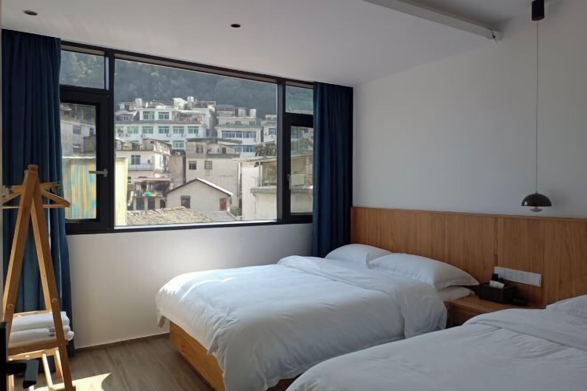 Habitación de hotel con dos camas, cabecera de madera, mesita de noche y un ventanal con vistas a edificios.